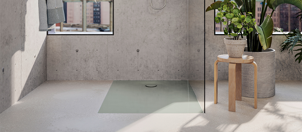 Vue intérieure d'une salle de bain moderne de style industriel, principalement en béton brut clair (murs et sol). Un receveur de douche encastré, de couleur gris-vert clair et affleurant le sol, est visible au centre, délimité par une paroi de douche en verre transparent. À droite, de grandes plantes vertes en pot sont posées sur le sol et sur un petit tabouret en bois clair. Des serviettes grises sont accrochées à gauche. La lumière du soleil entre par une fenêtre.